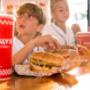 A red Five Guys soft drink cup is the focus in the foreground, as children enjoy their meals in the background. A red Five Guys soft drink cup is the focus in the foreground, as children enjoy their meals in the background.