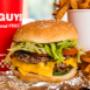 A close-up photograph of a Five Guys cheeseburger, soft drink in red Five Guys cup, and regular order of fries sitting on a table inside a Five Guys restaurant. A close-up photograph of a Five Guys cheeseburger, soft drink in red Five Guys cup, and regular order of fries sitting on a table inside a Five Guys restaurant.