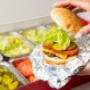 A close-up photo of a person putting the top bun of their cheeseburger in front of a Five Guys catering box of toppings. A close-up photo of a person putting the top bun of their cheeseburger in front of a Five Guys catering box of toppings.