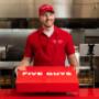 A Five Guys employee holds the red Five Guys catering box at the counter. A Five Guys employee holds the red Five Guys catering box at the counter.