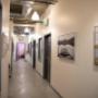Well-lit hallway with polished concrete floors, exposed ceilings, and framed art lining the walls. Leads to individual studio rooms, ideal for gallery-style displays. Well-lit hallway with polished concrete floors, exposed ceilings, and framed art lining the walls. Leads to individual studio rooms, ideal for gallery-style displays.
